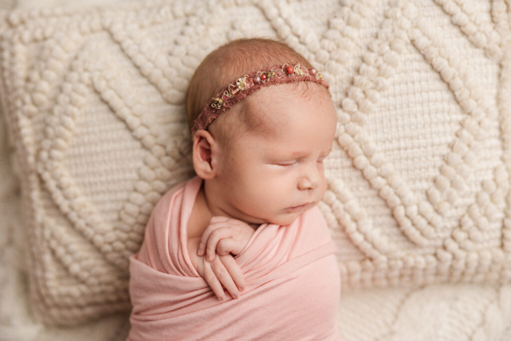 Newborn baby girl in studio. Baby is wrapped in a pink wrap with a maroon headband.