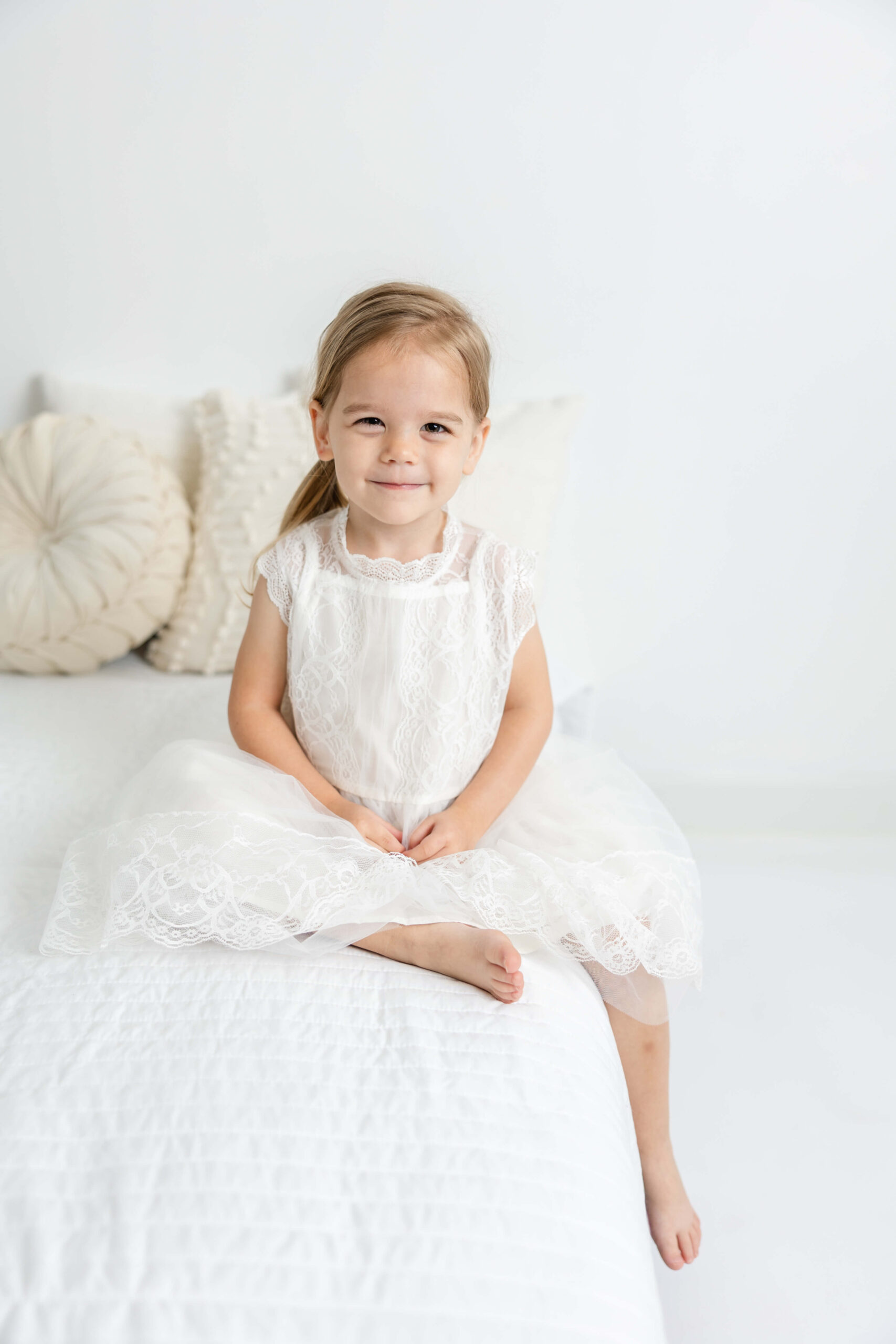 Two year old in white dress from client closet, sitting on bed during child portrait session.