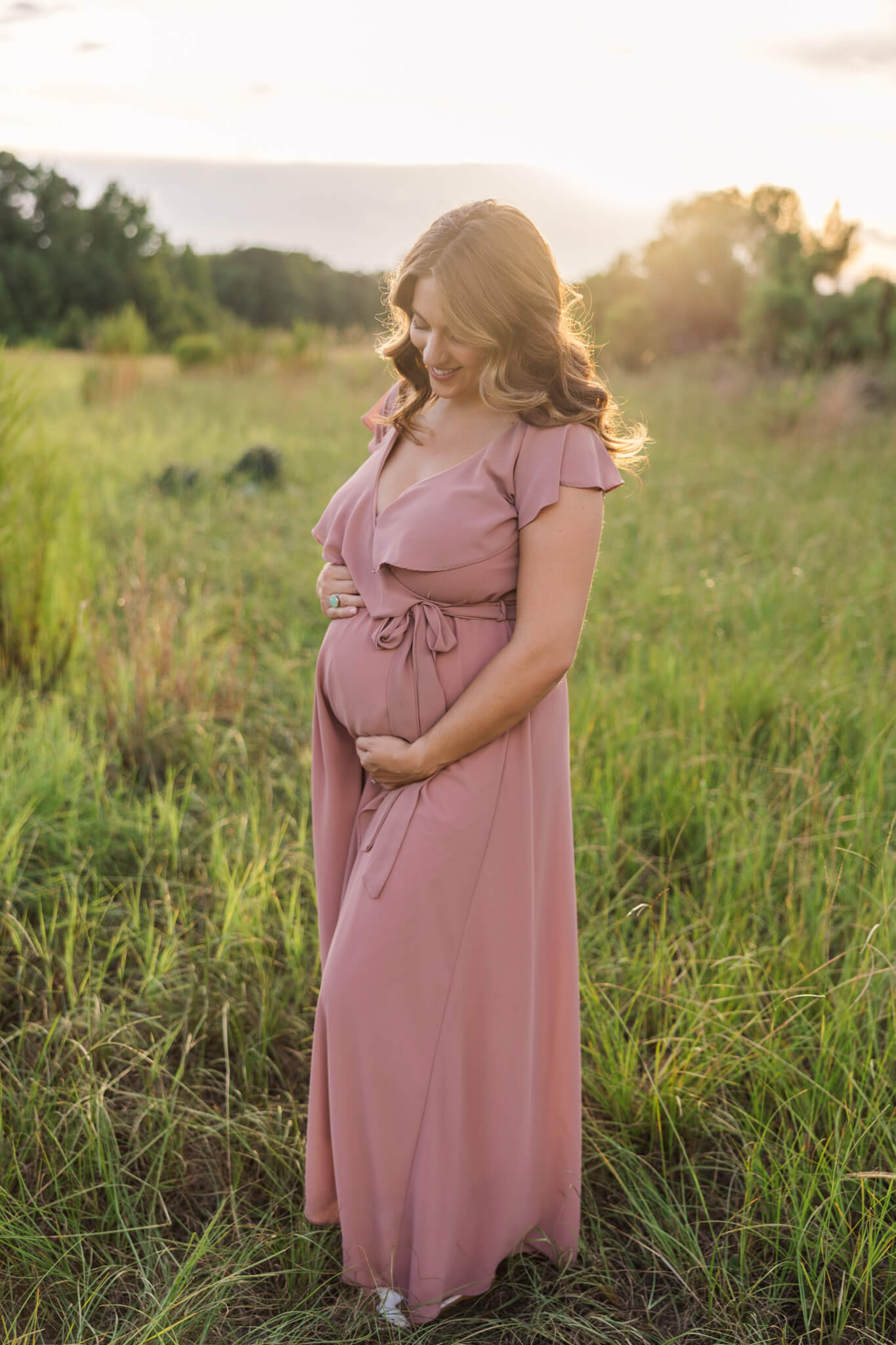 pregnant woman in pink maternity gown smiling at her growing bump at sunset in a field The Retreat Spa