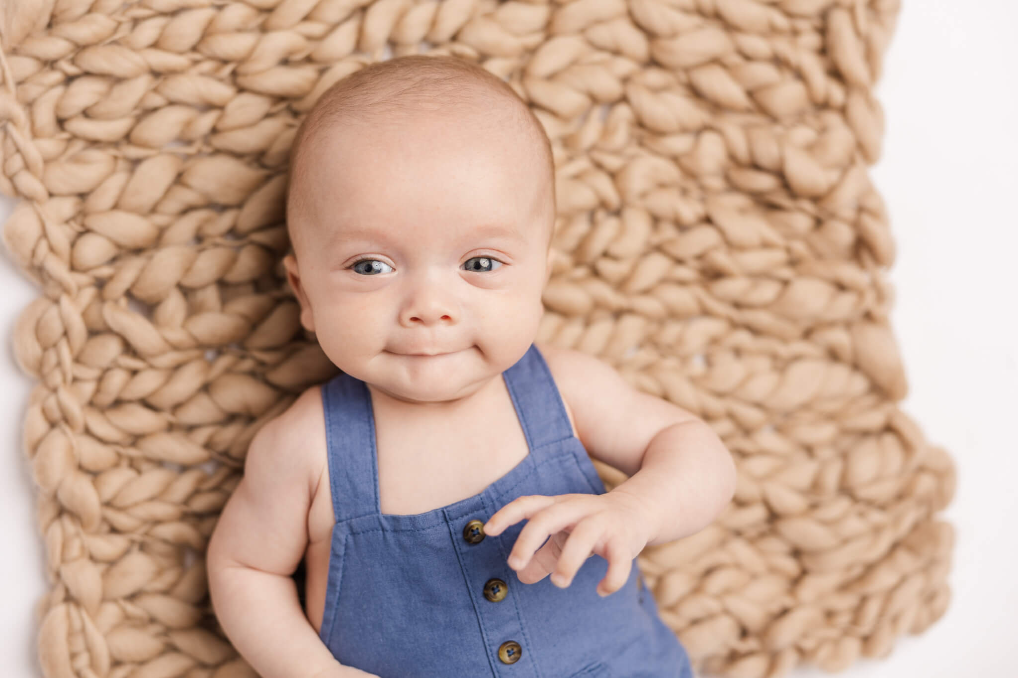 baby in blue overalls smirking while laying on a brown blanket