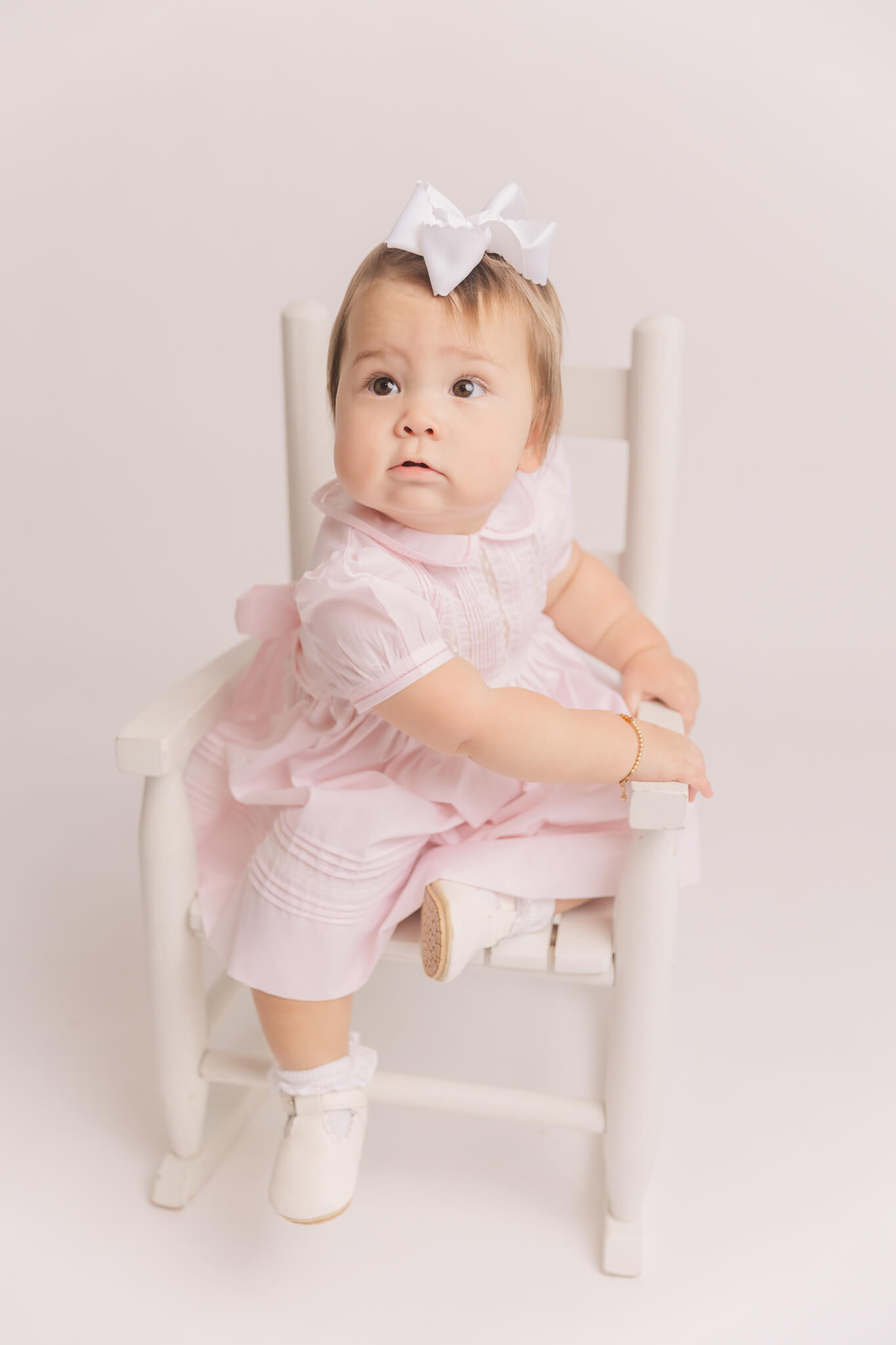 Little Girl in pink dress and white bow sitting in a white rocking chair Little Bear Smocks