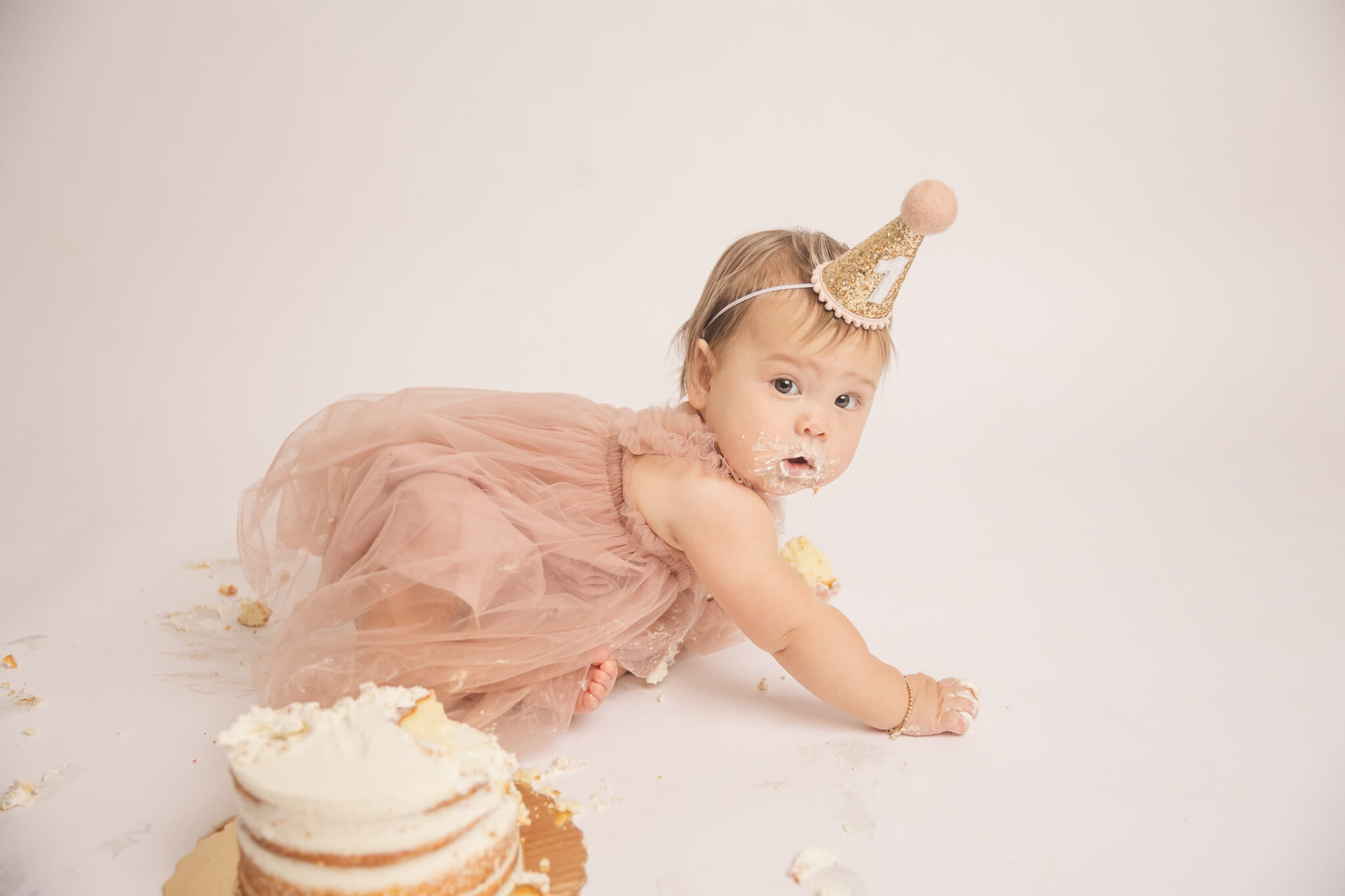 little girl in tulle pink dress and birthday hat crawling away from her cake