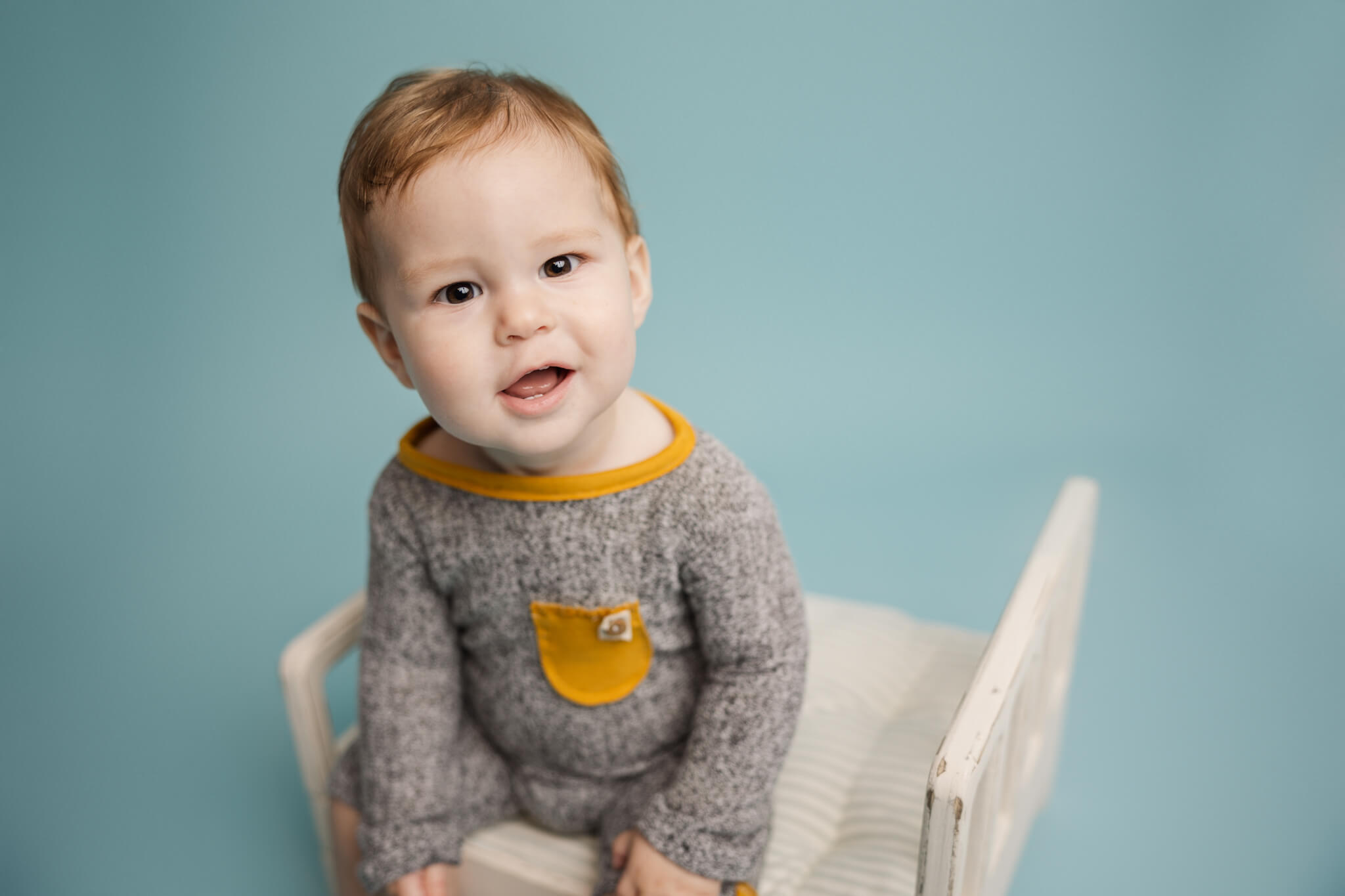 little boy in grey and yellow onesie sitting on a white wooden bed