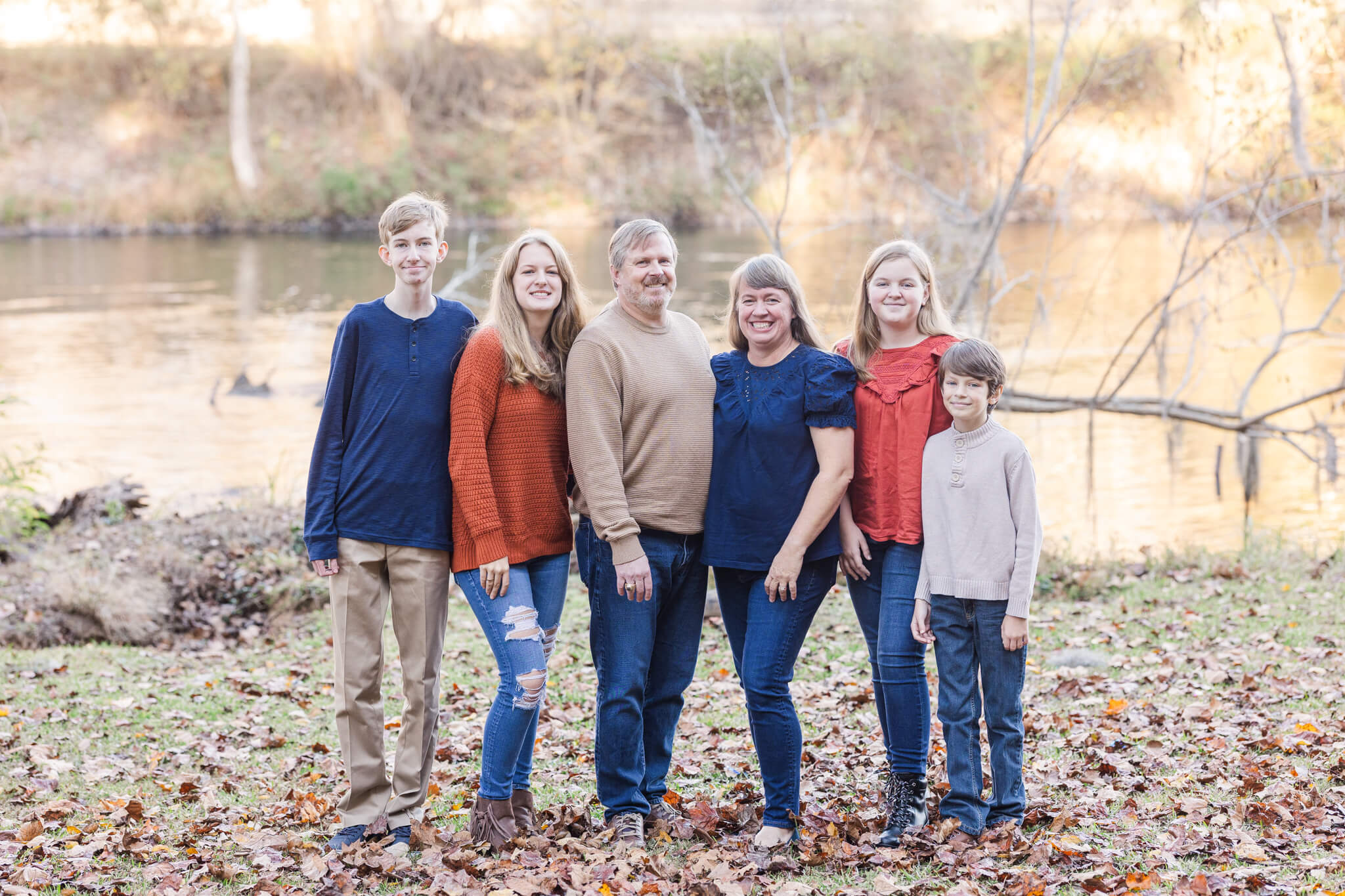 Family of six in blue, reds, and neutrals smiling near a lake Uptown Cheapskate Evans GA