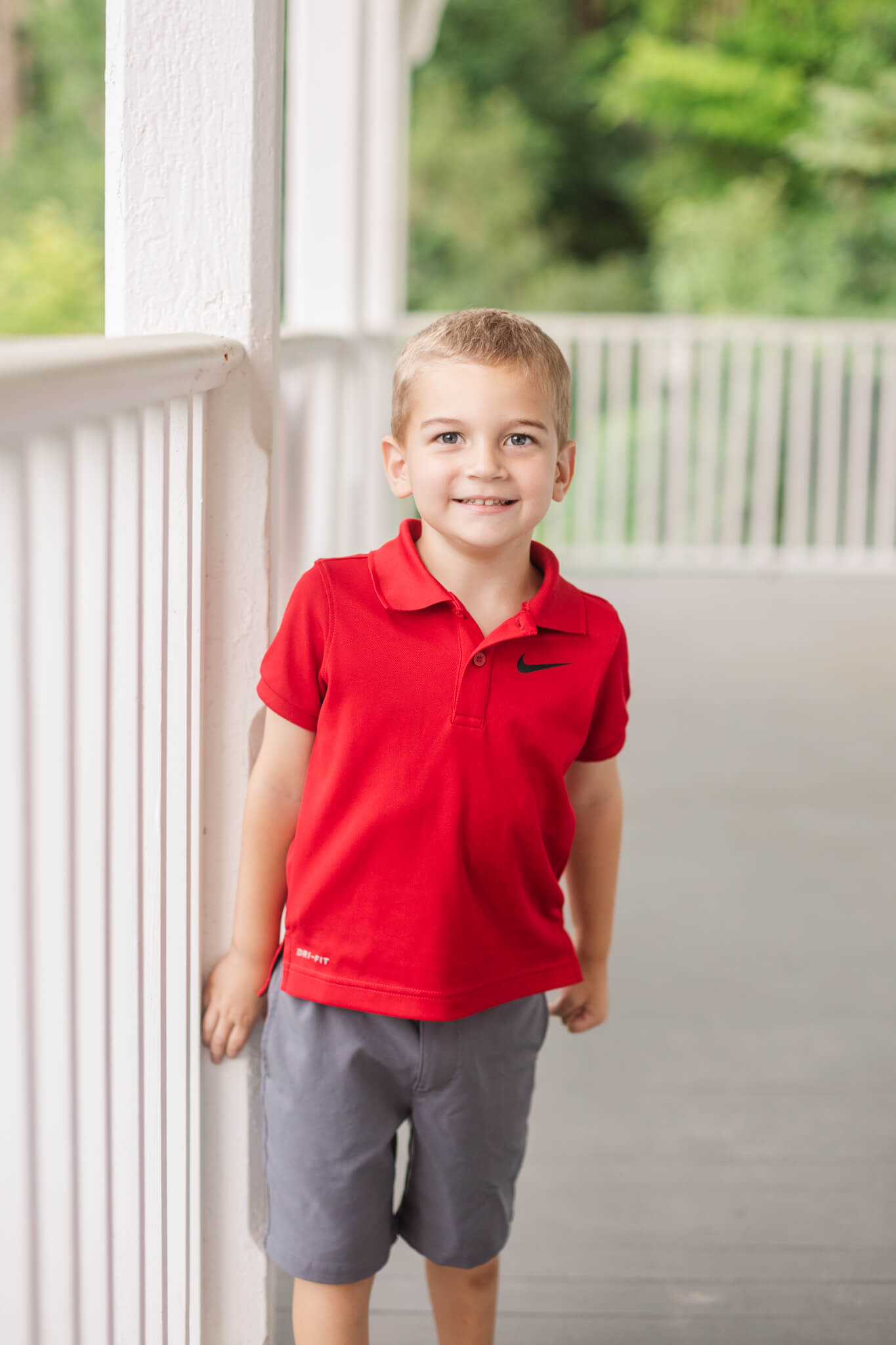 Little boy in red shirt and grey shorts smiling on a porch Southern Smiles Pediatric Dentistry