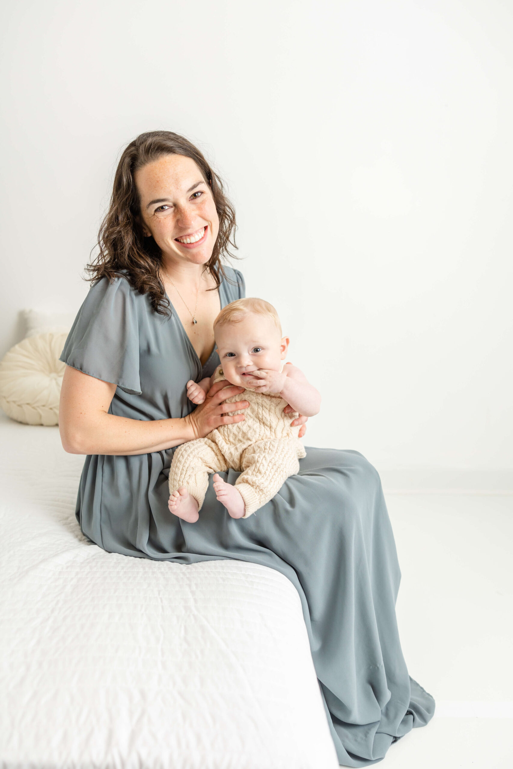Captured mom holding baby boy while sitting on edge of bed during their studio motherhood session. 
