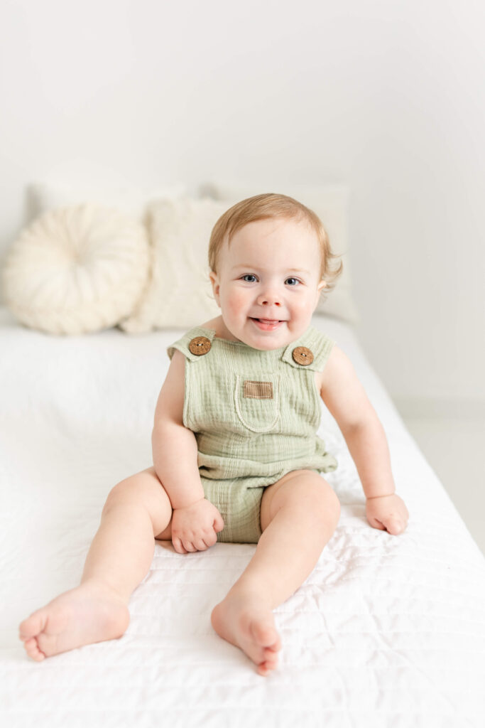 1 year old boy, in light green jumper, sitting up on a white bed. 
