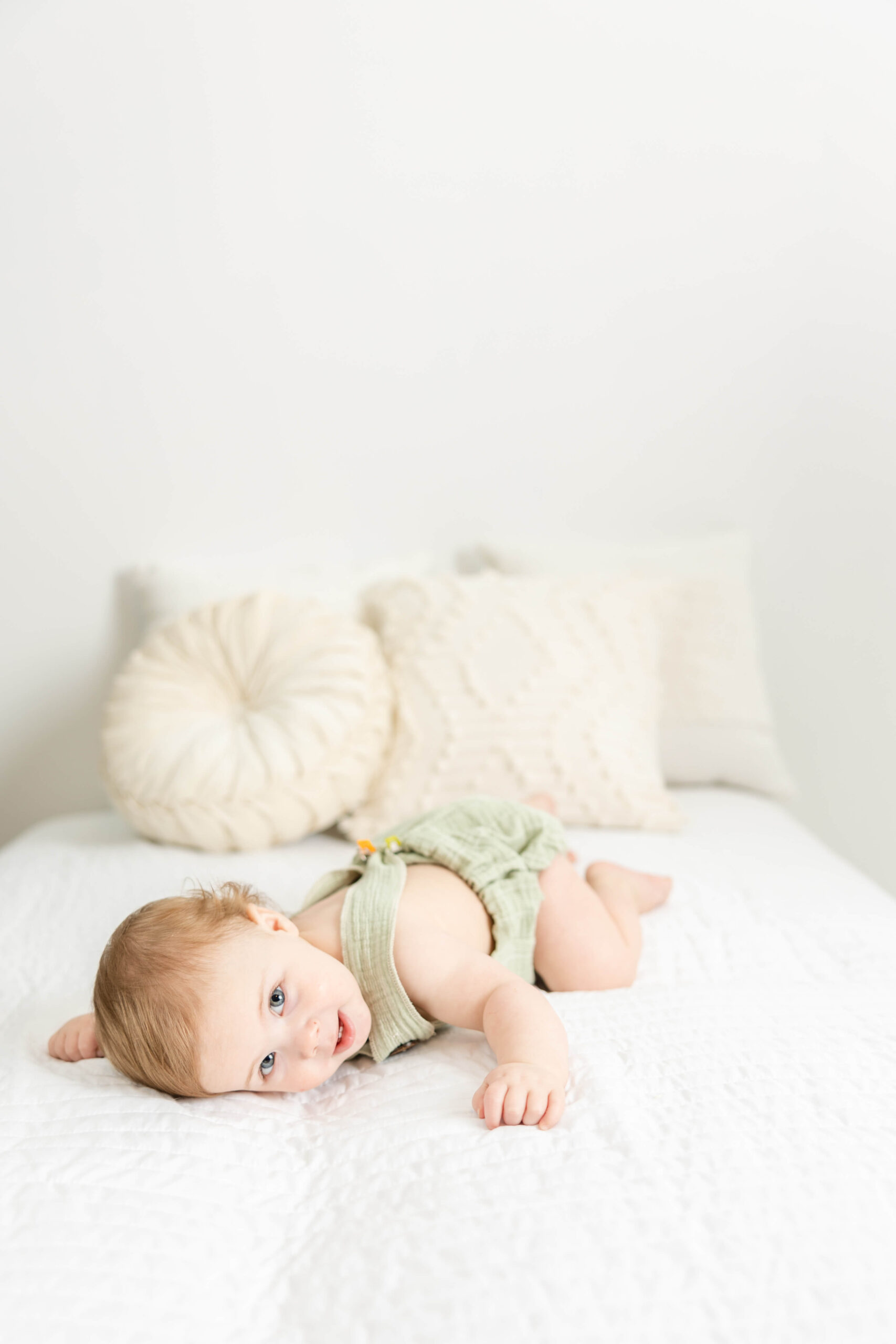 1 year old boy in green romper laying on a white bed during his milestone session. 