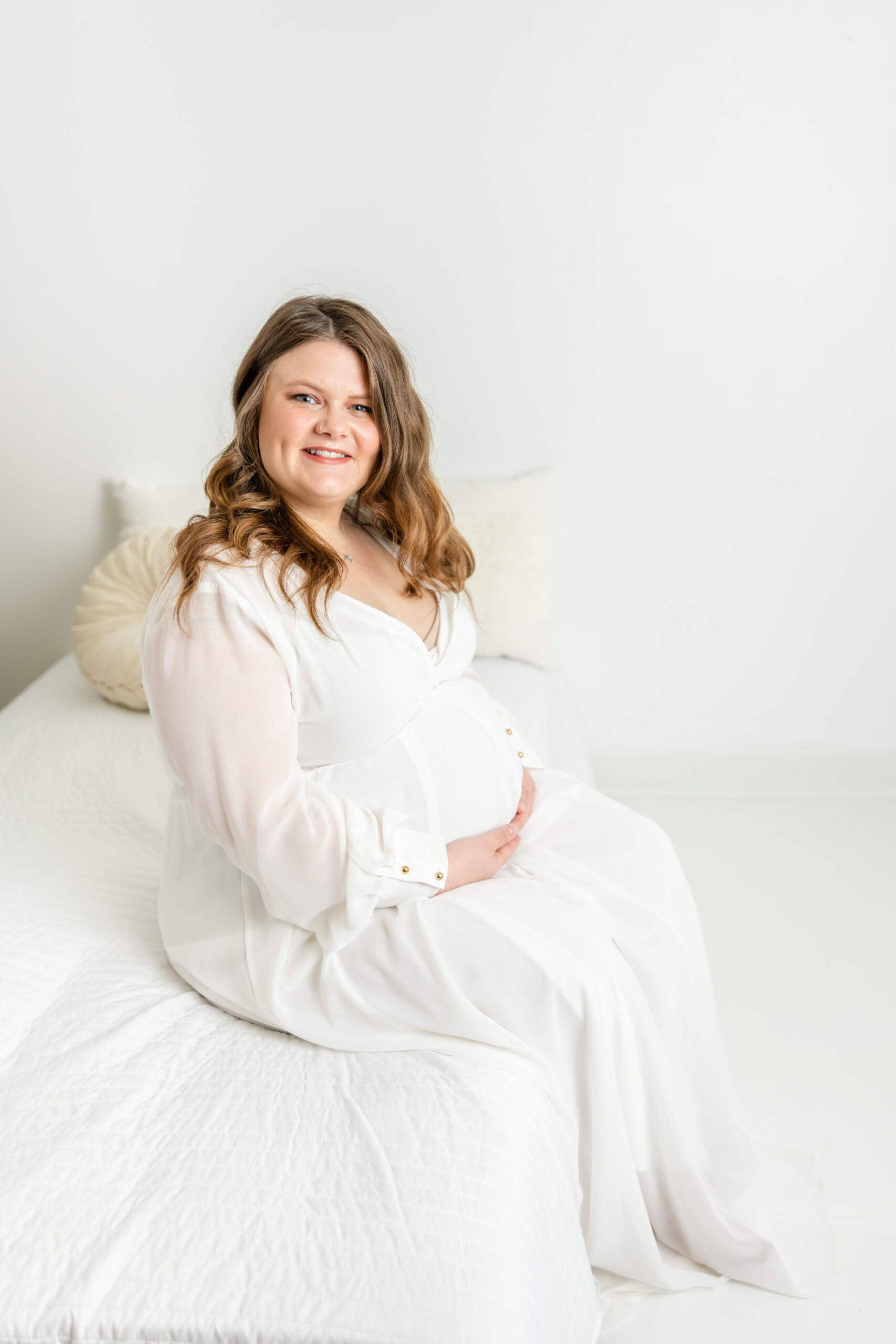 Expecting mom sitting on white bed in full length white dress, during studio maternity session. 