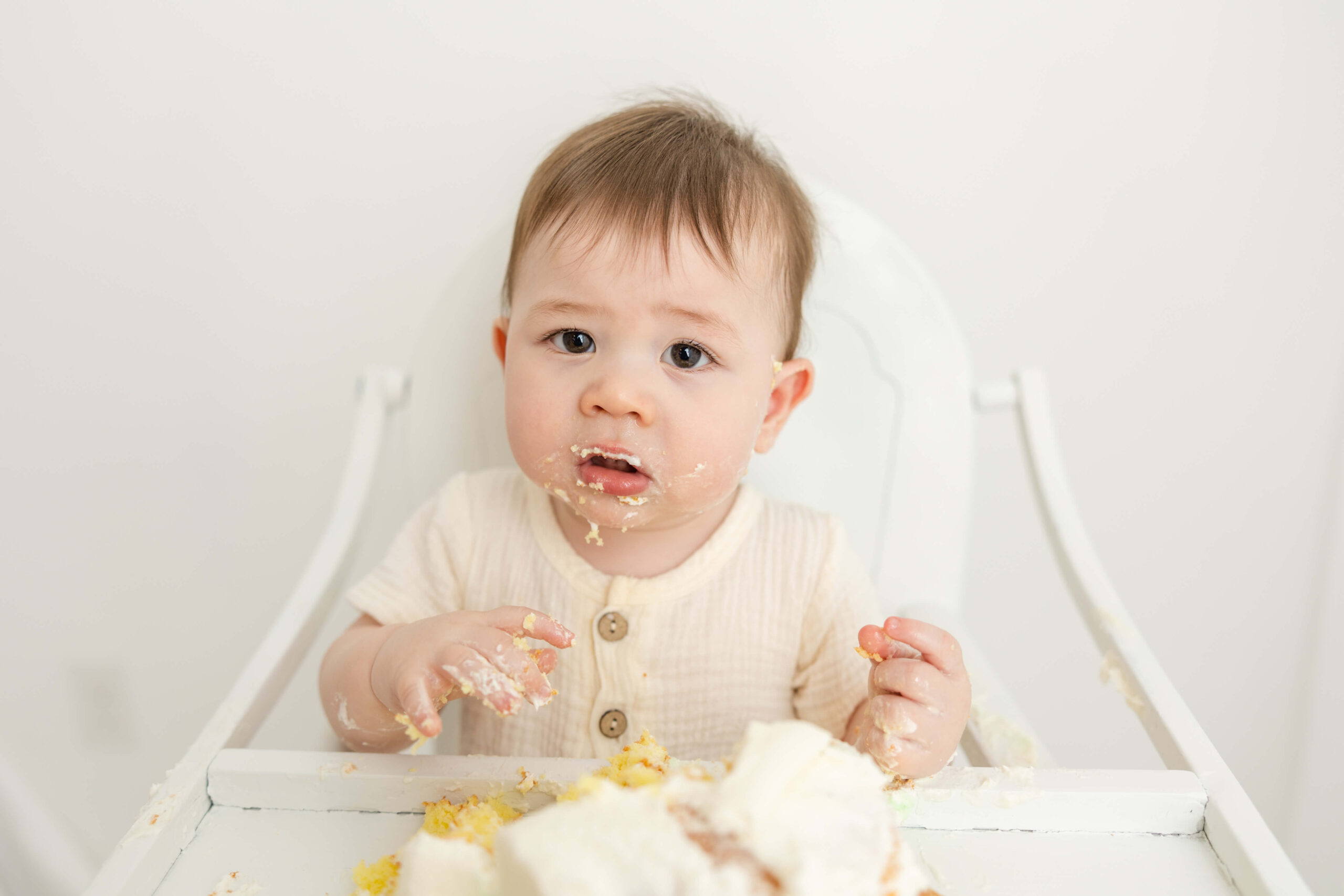 Little boy in white high chair with cake on his face from his cake smash session. 