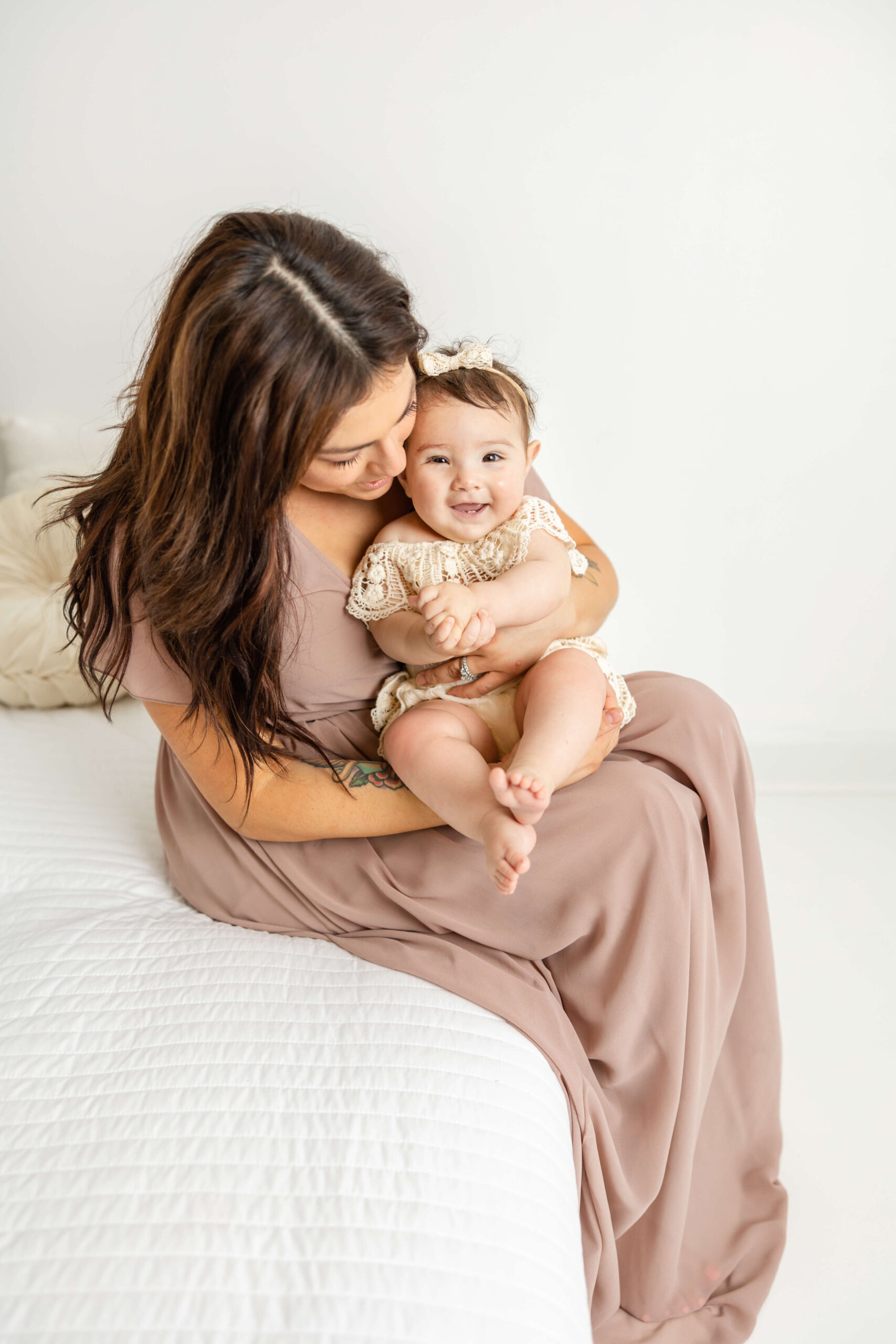 At their motherhood session, mom in a mauve dress and her little girl in a cream romper are snuggling together as they sit on the edge of a bed. 