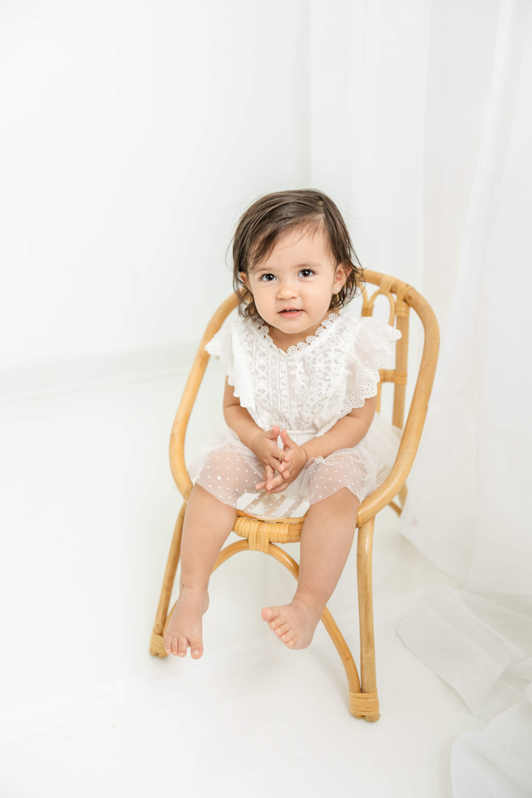 Little girl in white romper sitting in a wicker brown chair.