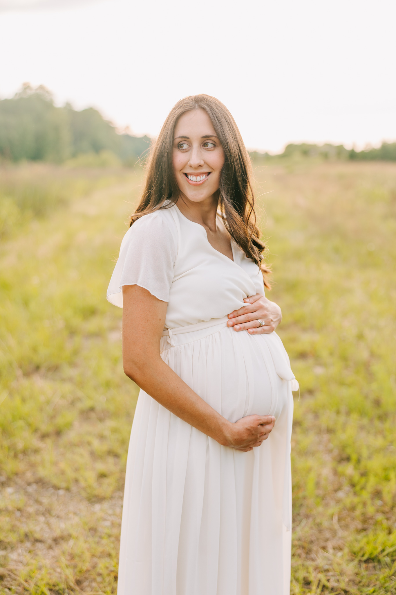 expecting mom holding her belly during her maternity session. Mom is wearing a white dress.